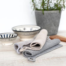 Lade das Bild in den Galerie-Viewer, Handgestrickte Spueltuecher in grau und beige liegen auf hellem Holz mit Keramik-Schalen im Hintergrund