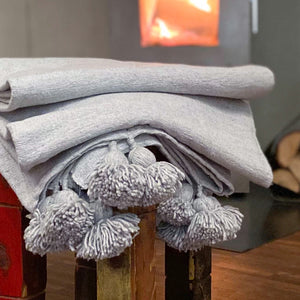 Eine handgewebte graue Pompom-Decke liegt auf einem Hocker vor einem Kaminofen.