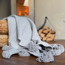 Load image into Gallery viewer, Eine handgewebte graue Pompom-Decke aus Baumwolle liegt auf einem Hocker vor einem Kaminofen.

