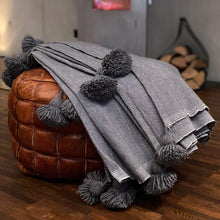 Load image into Gallery viewer, Eine handgewebte graue Pompom-Decke aus Baumwolle liegt auf einem Pouf vor einem Kaminofen.
