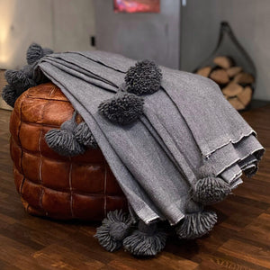 Eine handgewebte graue Pompom-Decke aus Baumwolle liegt auf einem Pouf vor einem Kaminofen.