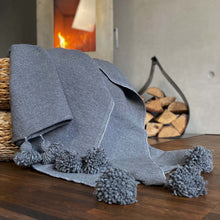 Lade das Bild in den Galerie-Viewer, Eine handgewebte graue Pompom-Decke aus Baumwolle liegt auf einem Pouf vor einem Kaminofen.

