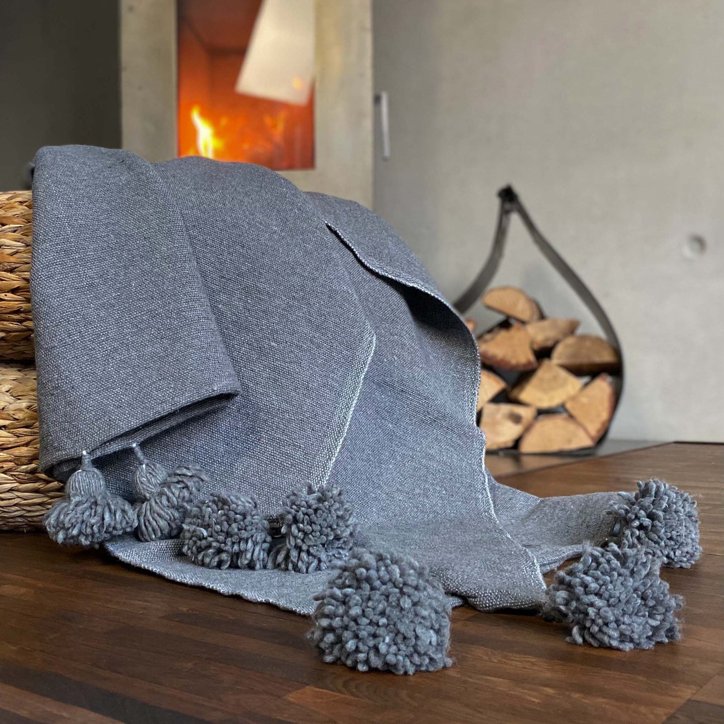 Eine handgewebte graue Pompom-Decke aus Baumwolle liegt auf einem Pouf vor einem Kaminofen.