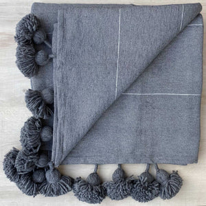 Graue Decke mit Bommeln liegt auf einer hellen Holz-Platte