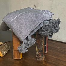 Load image into Gallery viewer, Eine handgewebte graue Pompom-Decke aus Baumwolle liegt auf einem Hocker vor einem Kaminofen.

