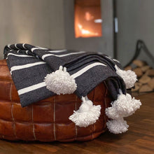 Load image into Gallery viewer, Eine handgewebte grau-weiß-gestreifte Pompom-Decke aus Baumwolle liegt auf einem Hocker vor einem Kaminofen.
