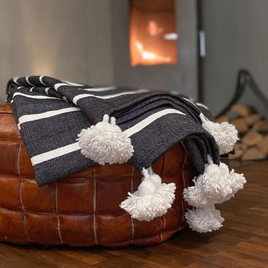 Eine handgewebte grau-weiß-gestreifte Pompom-Decke aus Baumwolle liegt auf einem Hocker vor einem Kaminofen.