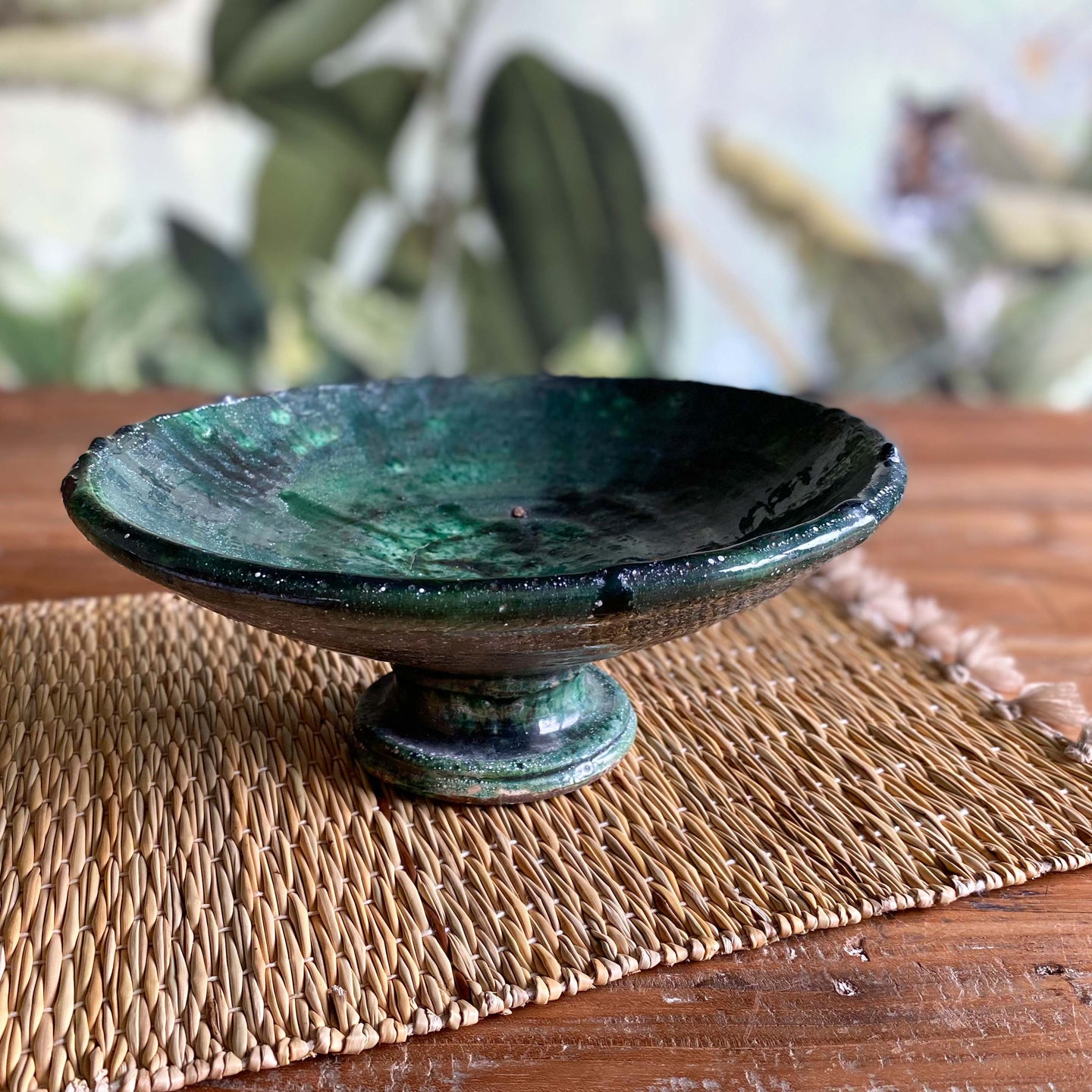 Eine marokkanische handgearbeitete grüne Keramik-Schale mit Fuß steht auf Vintage-Holztisch vor einer floralen Tapete